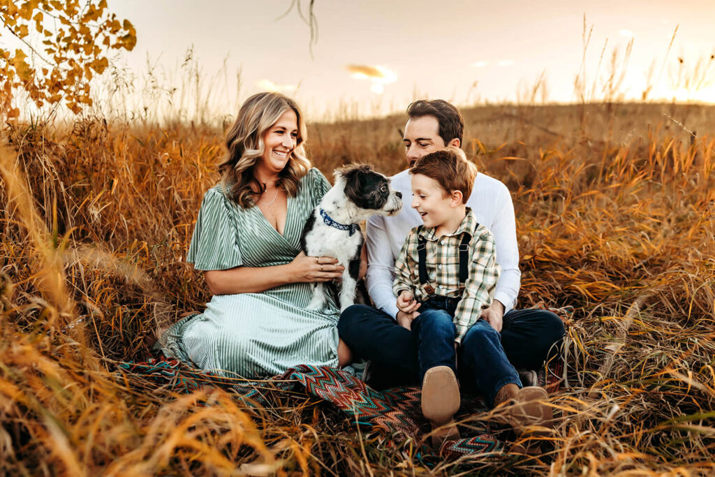 Mom, dad, young son and small dog sitting together in a Berthoud field laughing together. 