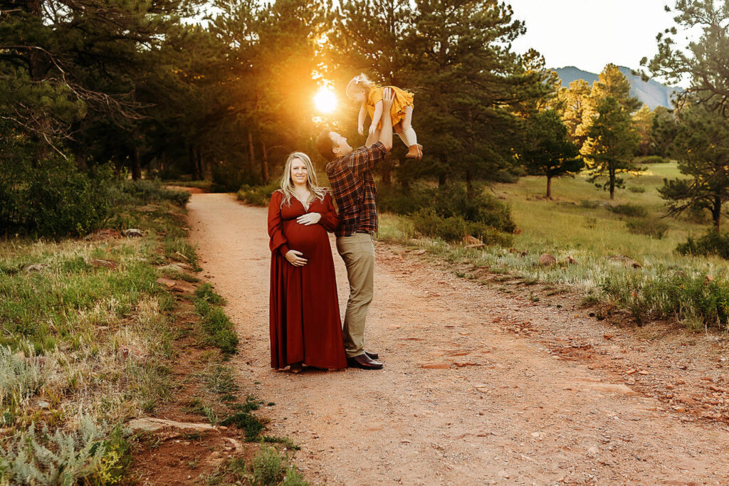 Expecting mom looking and smiling as dad throws toddler daughter in the air in the Boulder sunset. 