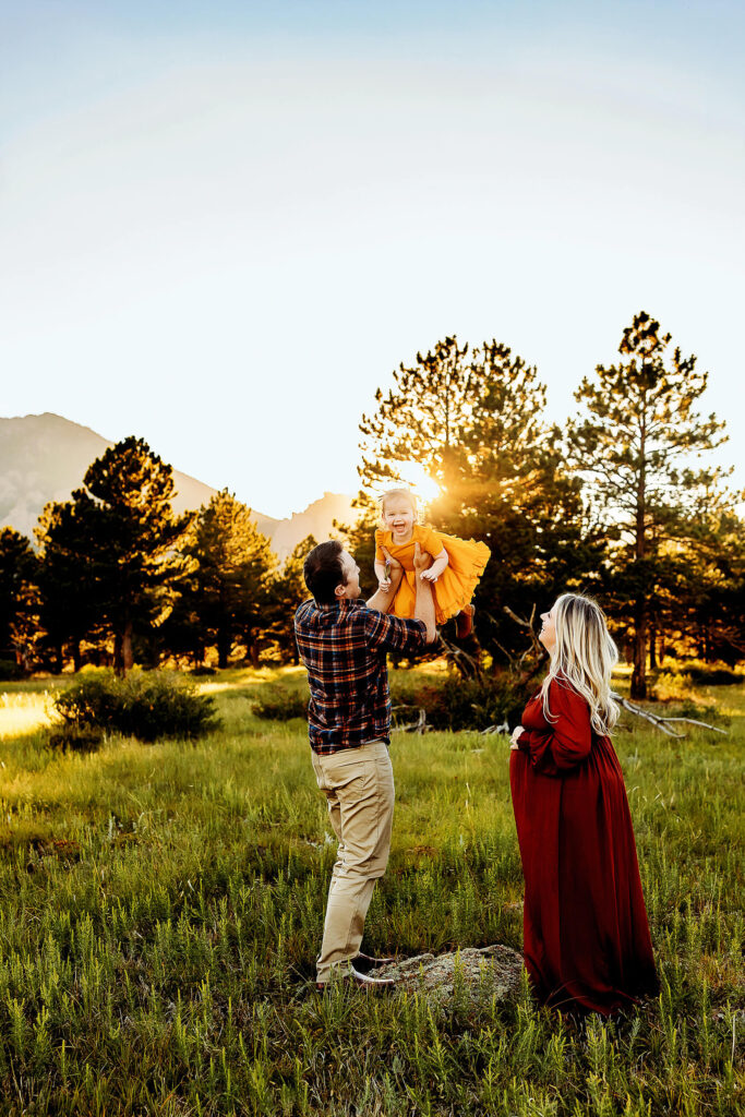 Dad throwing toddler daughter up in the air as pregnant mom watches and smiles. 