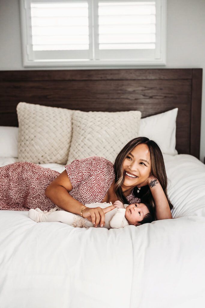 New mom laying with her baby daughter on the bed while she smiles at her husband.